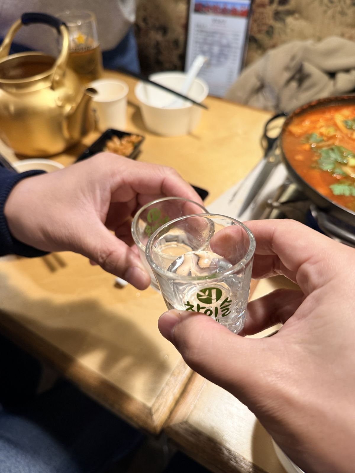 兩個燒酒杯乾杯,背景是辣鍋與金色茶壺